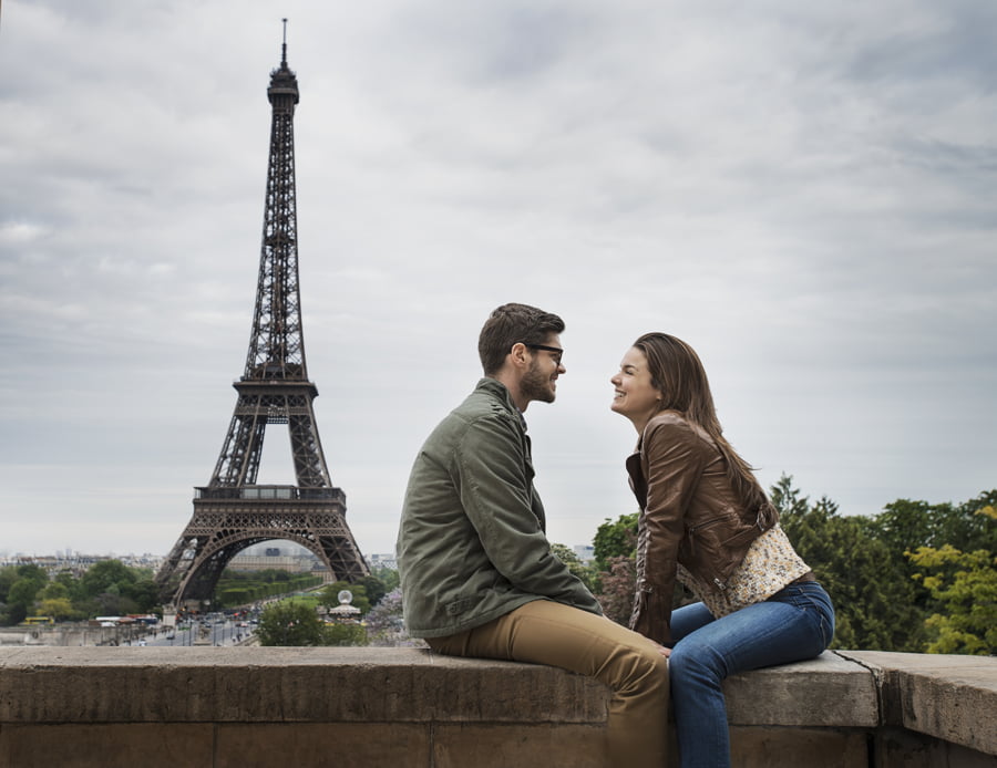 pareja sentada frente a frente en un muro de la ciudad de París con la Torre Eiffel al fondo.