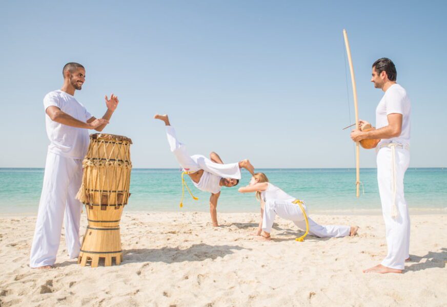 Equipo de capoeira entrenando en la playa - Atletas de artes marciales realizando acrobacias mientras dos hombres tocan música