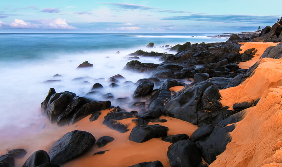 Larga exposición de olas rompiendo en una playa de arena anaranjada al atardecer.